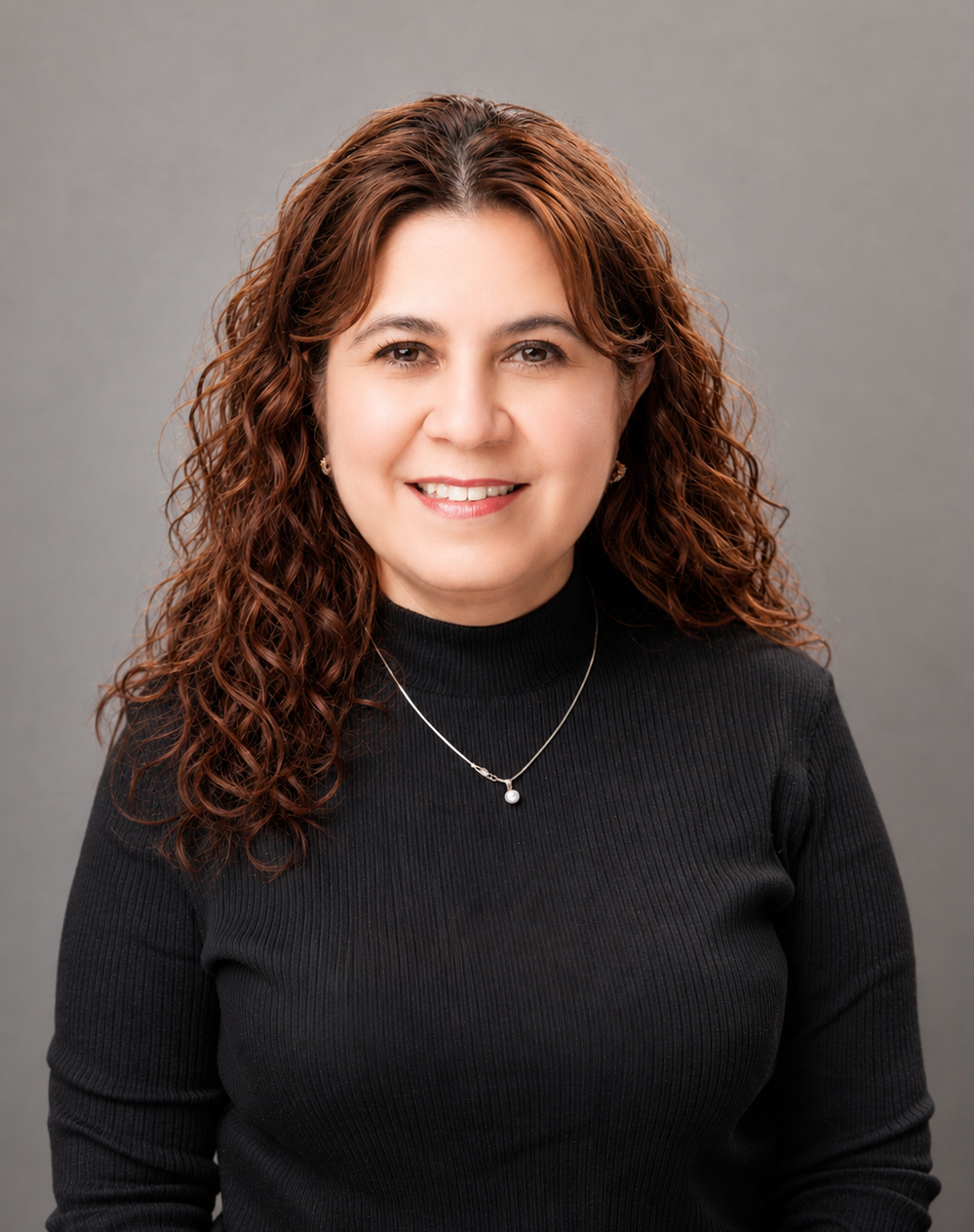 Woman with long curly brown hair wearing a black ribbed sweater and a necklace, smiles at the camera in front of a plain gray background.