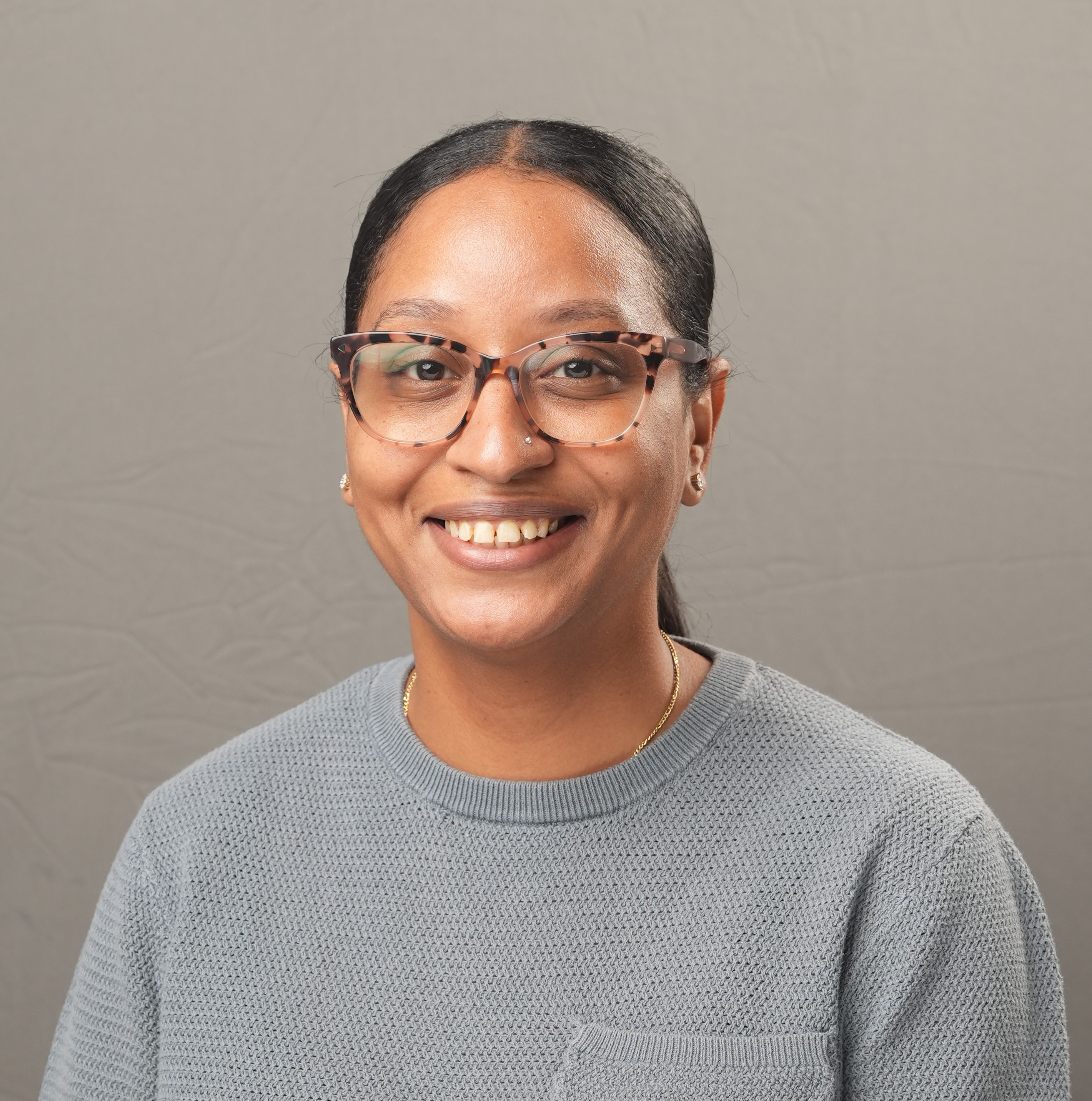 A woman with glasses and long dark hair pulled back, wearing a grey sweater, smiles at the camera against a plain grey background.