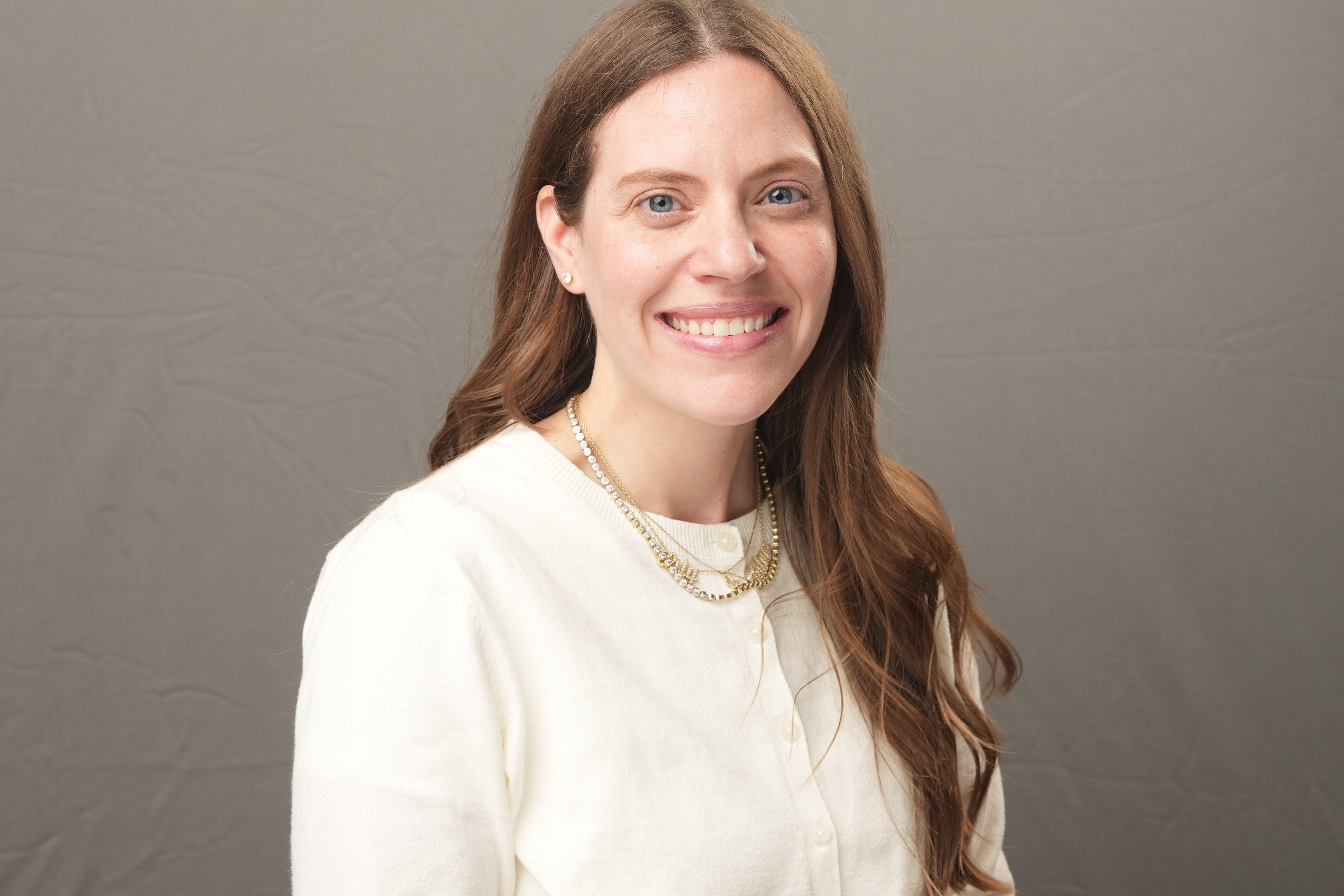 Woman with long brown hair wearing a white top and gold necklace, smiling and posing in front of a plain gray background.
