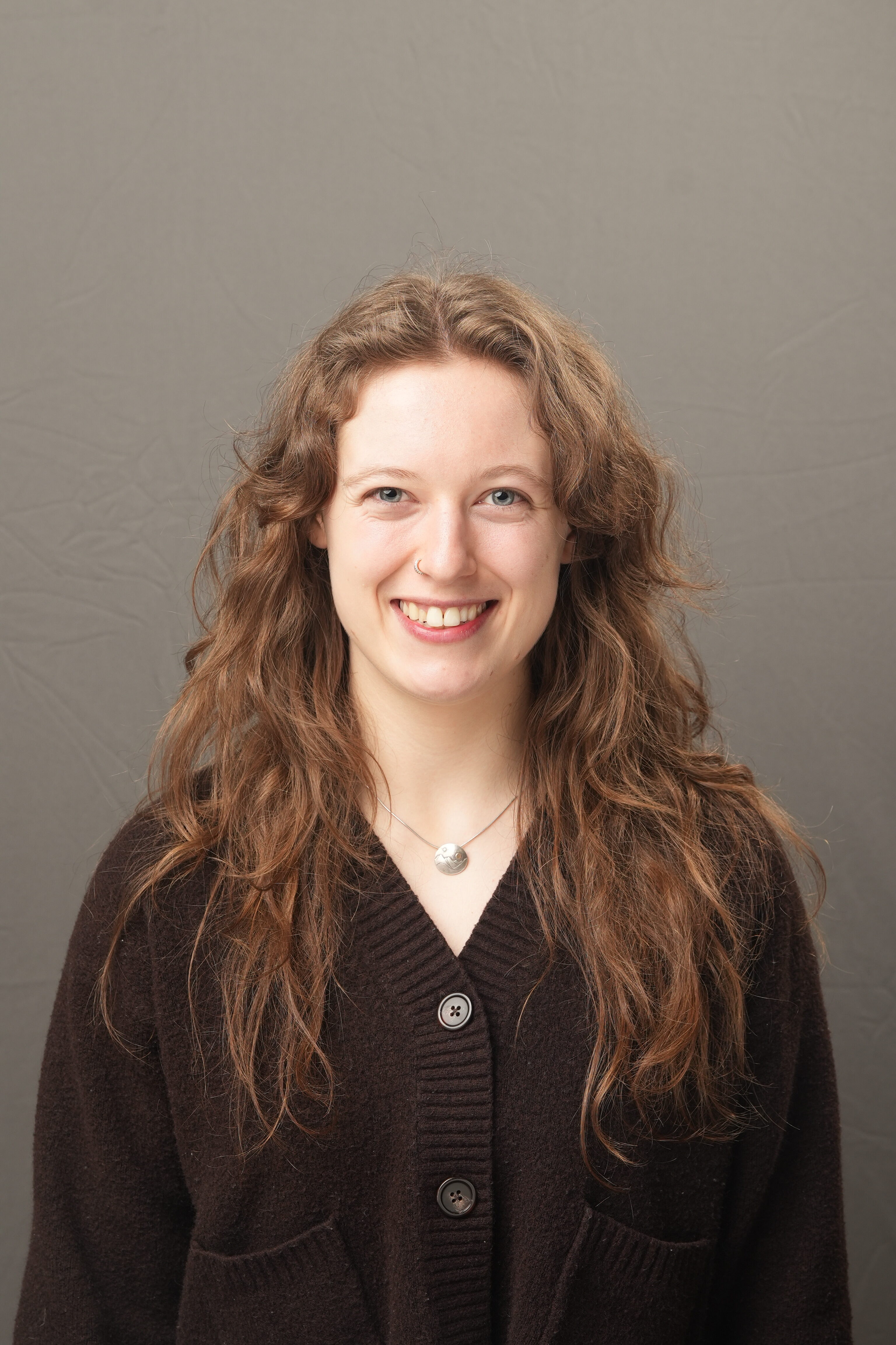 A young woman with long wavy brown hair, wearing a dark sweater and necklace, smiles at the camera against a plain gray background.