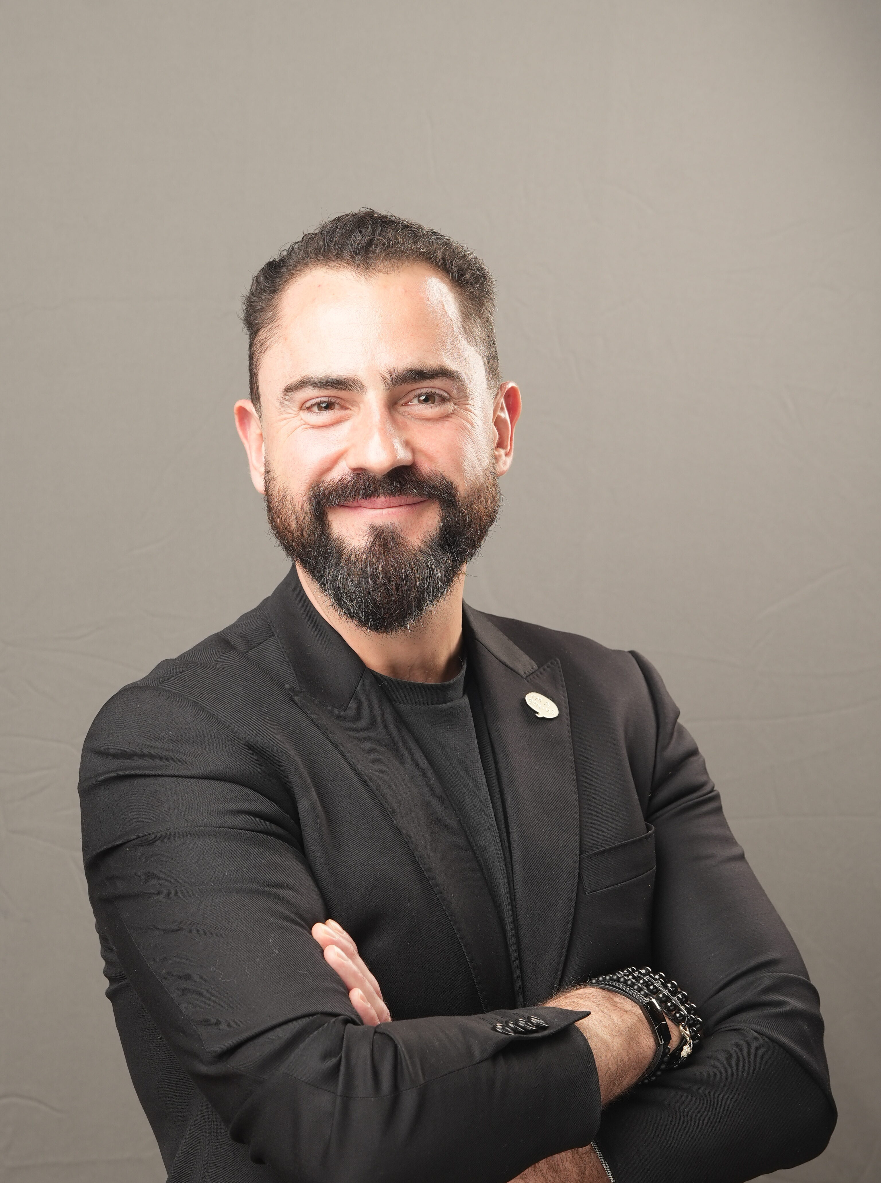 Man with dark hair and beard, wearing a black suit and black shirt, stands with arms crossed and smiling against a plain gray background.