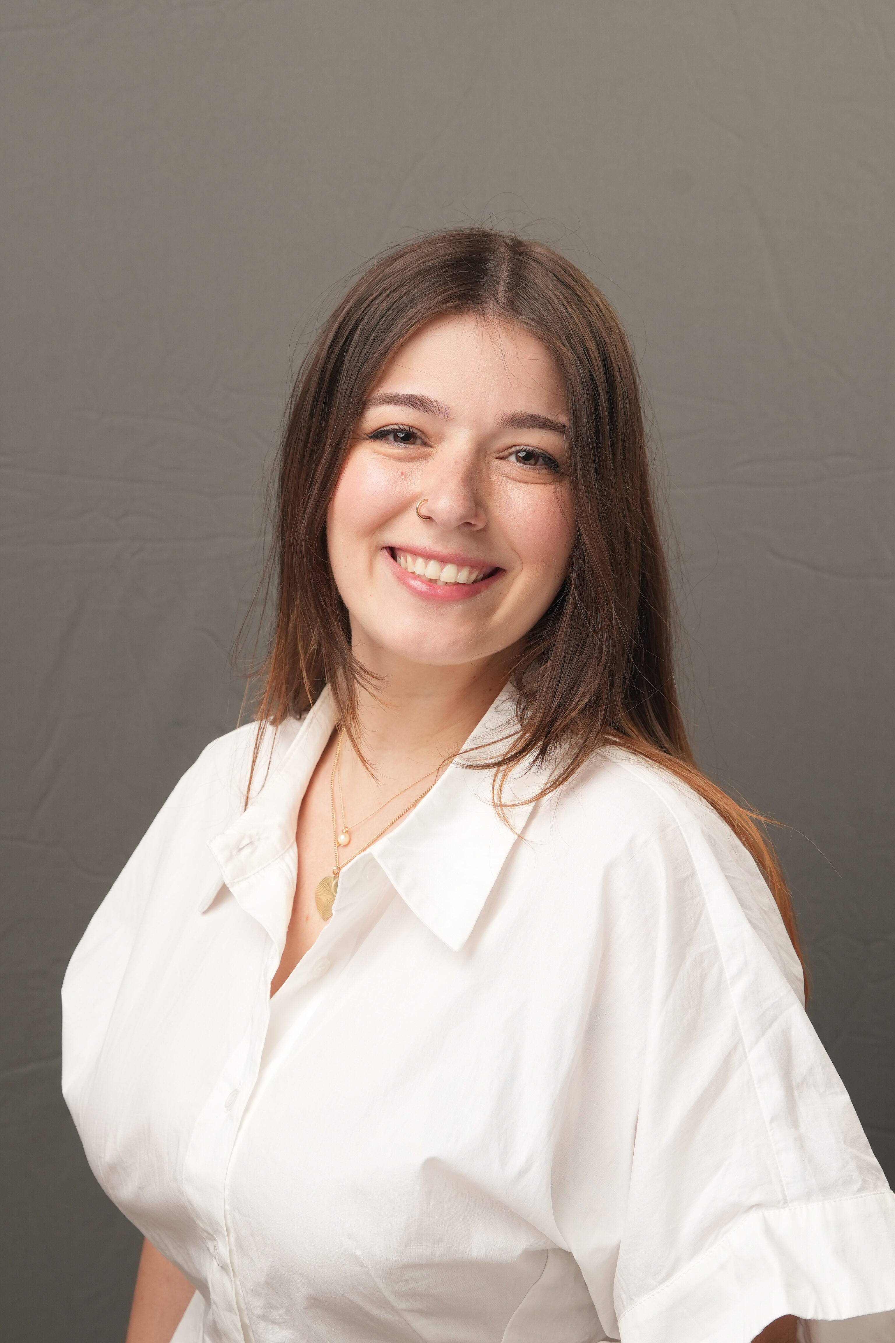 Allison Schwartz, a woman with long brown hair, wearing a white collared shirt and gold necklace, smiles in front of a plain gray background.