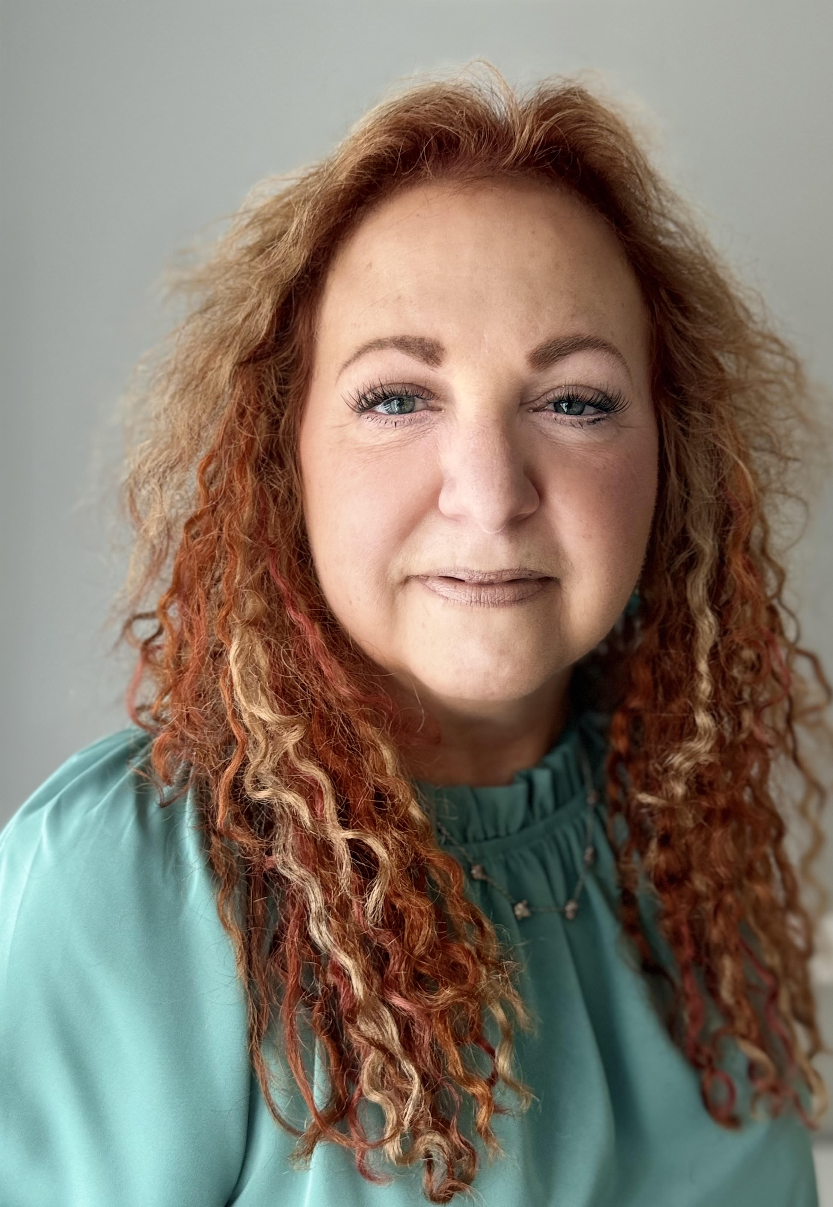 Woman with curly red and blonde hair, light skin, and blue eyes, wearing a teal blouse, looking at the camera with a neutral expression against a plain background.