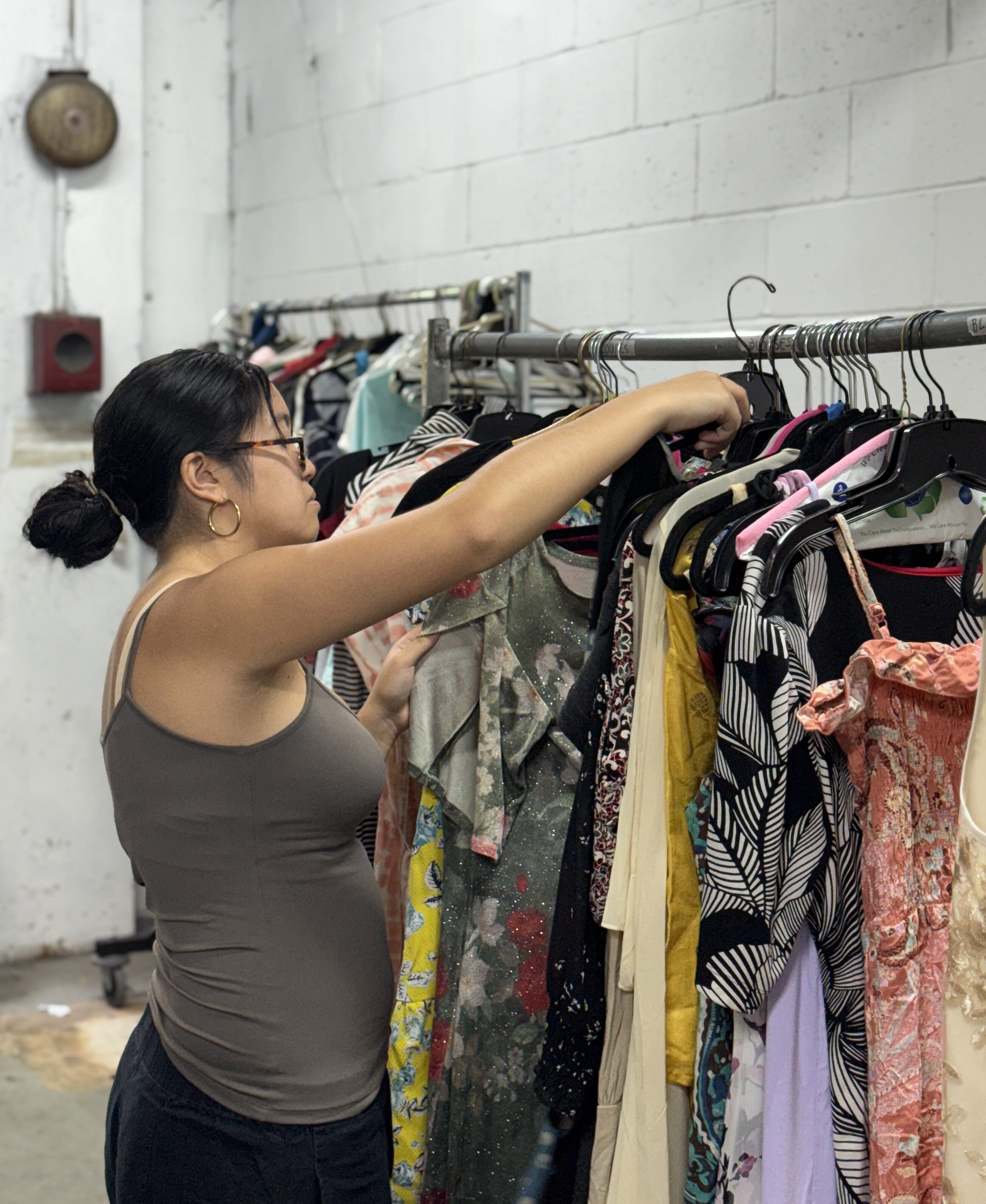 A woman with glasses browses through a rack of professional attire in an industrial-style room with white brick walls, selecting options that could support job seekers from low-income families.