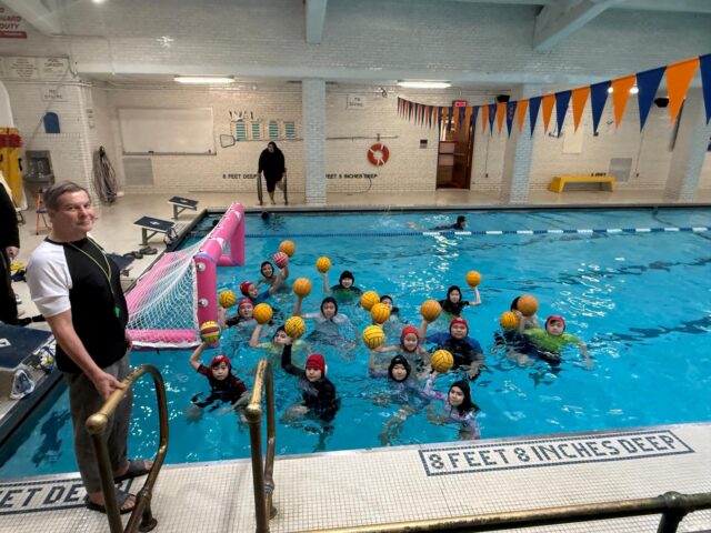 A group of children in swim caps hold yellow water polo balls in a pool at the Commonpoint Water Polo Club, with an adult by the ladder and another adult standing poolside near a goal during tryouts.