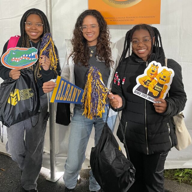 Three young women stand together holding college-themed props and gift bags, smiling at the camera in front of a white tent backdrop.
