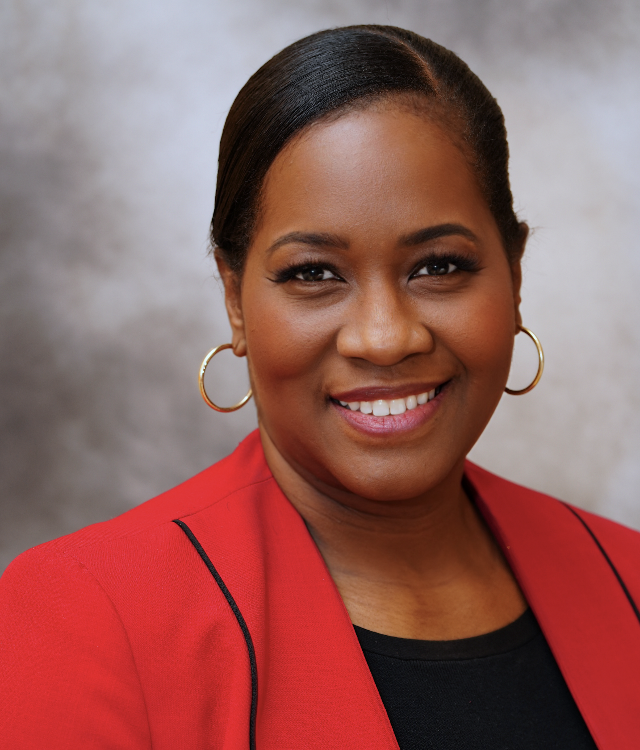 A woman in a red blazer and gold hoop earrings smiles at the camera against a blurred gray background.