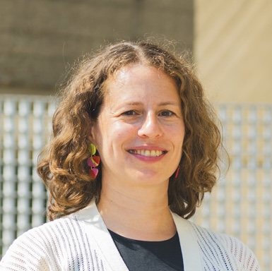 A woman with curly brown hair smiles outdoors, wearing a white sweater, black top, and colorful earrings.