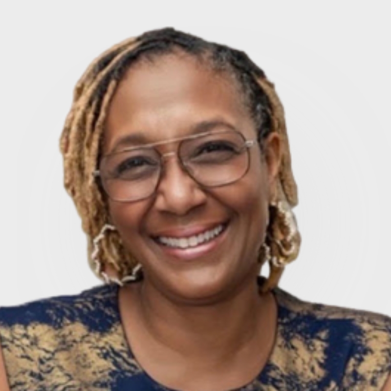 Smiling Dr. Lisa DeRoché, wearing glasses and light brown dreadlocks, is pictured in a patterned blue and gold top against a plain light background.