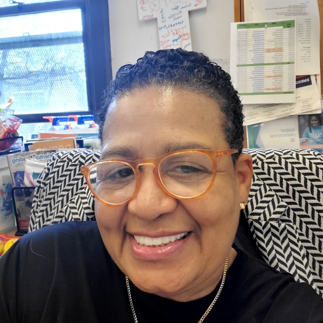 Norma Galloway, a person with short curly hair and orange glasses, is smiling while seated indoors with office papers, books, and a window in the background.
