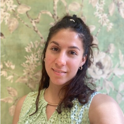 Woman with long dark hair and a sleeveless green top stands in front of a green floral-patterned background, smiling softly at the camera.