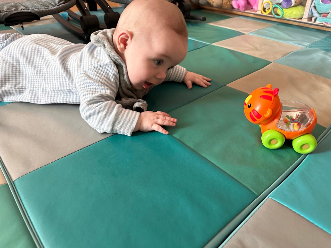 A baby in a striped shirt lies on a checkered floor mat, reaching toward a toy tiger with wheels.