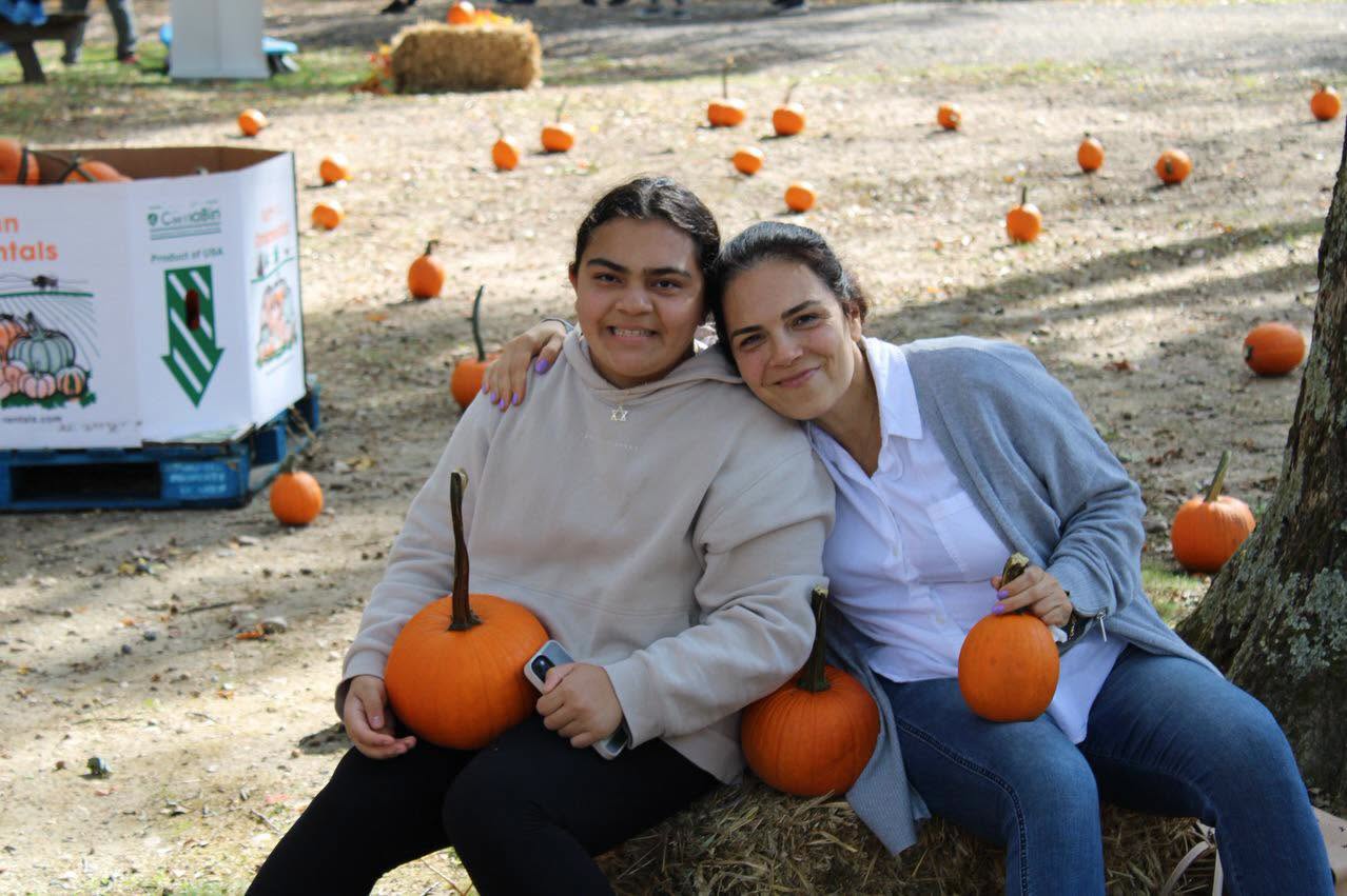Two people sit on a hay bale at an autumn pumpkin patch, each holding a small pumpkin.