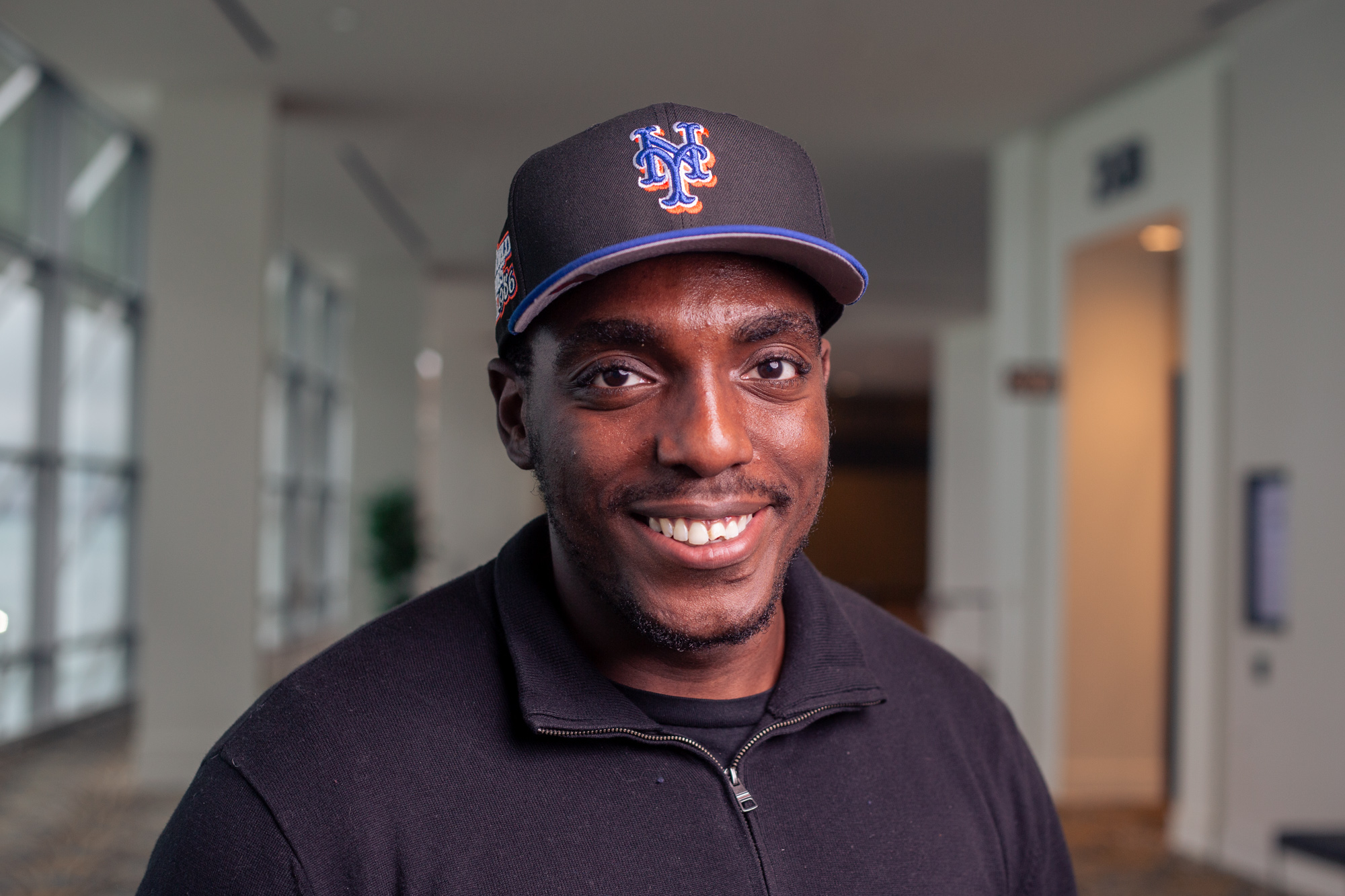 D’Andre Johnson wears a black New York Mets baseball cap and a black zip-up sweater while smiling at the camera.