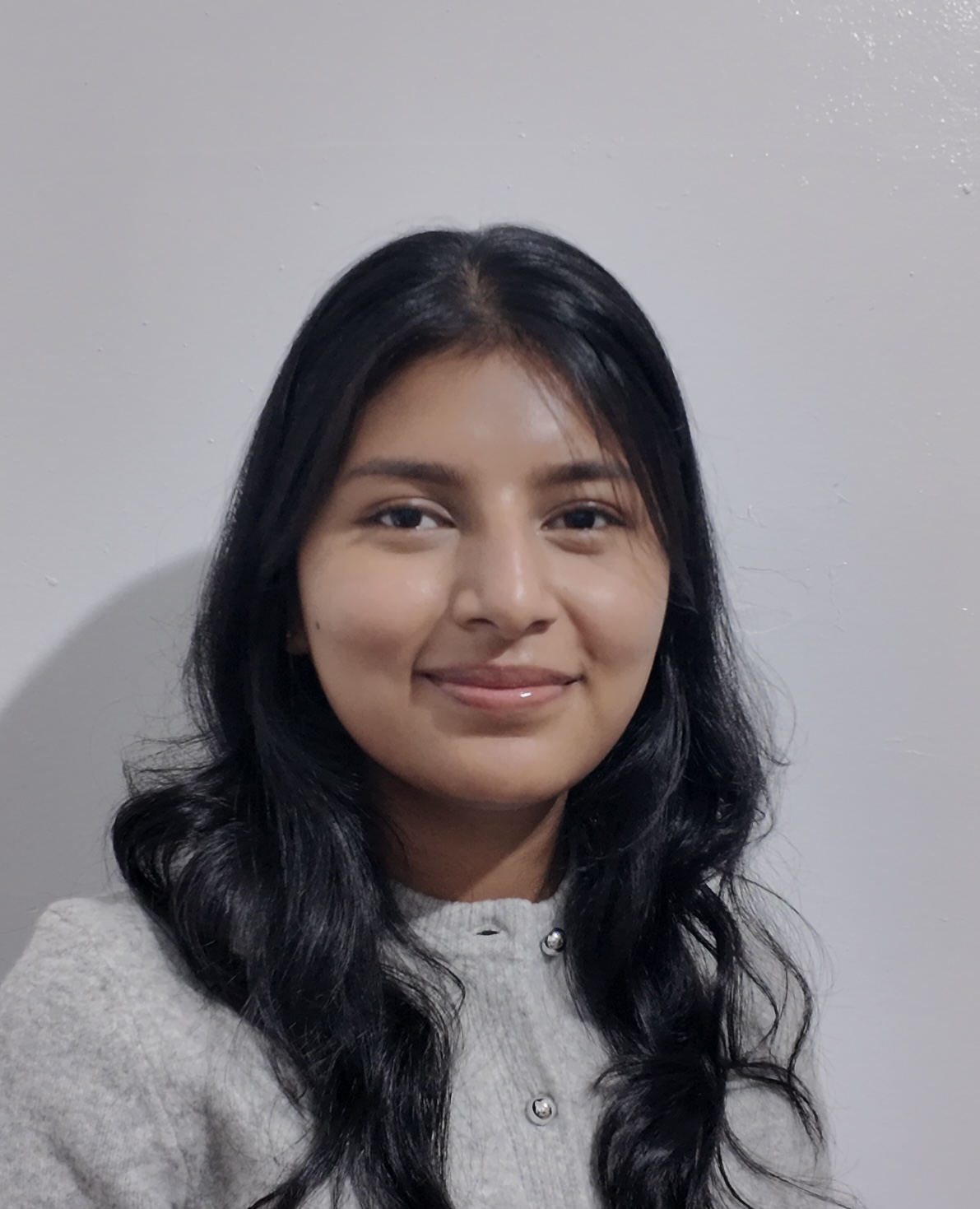 Portrait of a young woman with long dark hair smiling against a plain white wall.